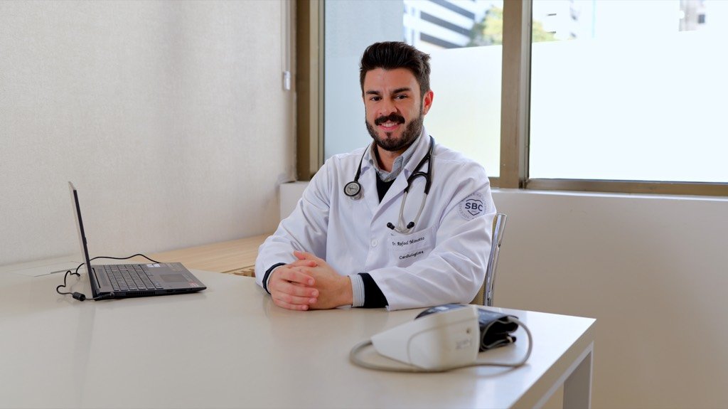 Cardiologista de jaleco e estetoscópio sentado à mesa do consultório, junto à janela.