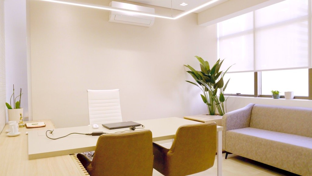 Consultório com mesa de atendimento, duas poltronas para acompanhantes e sofá junto à janela.