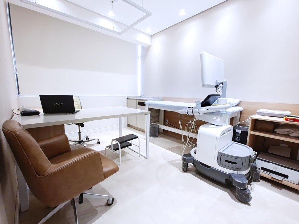 Sala ampla de diagnóstico com poltrona de couro, mesa de laudo e equipamento de exame.