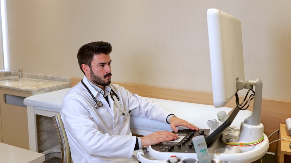 Cardiologista operando o painel do aparelho de ecocardiografia, sentado ao lado da maca.