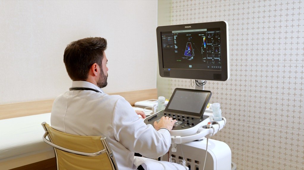 Cardiologista de costas diante do aparelho de ultrassom, com a imagem do coração em Doppler colorido na tela.