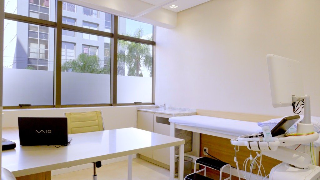 Sala de exame com maca articulada junto à janela e estação de trabalho do cardiologista.