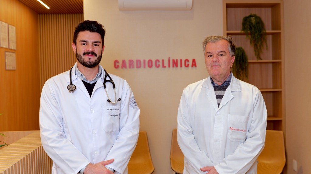 Dois cardiologistas de jaleco branco, lado a lado na recepção, com o nome Cardioclínica na parede ao fundo.